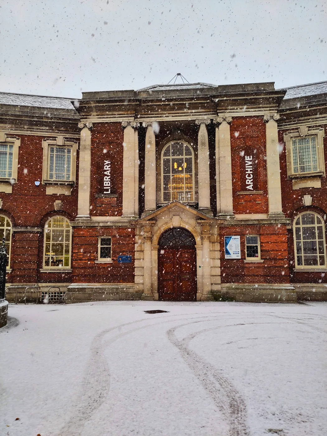 The red-brick front of York Explore Library and Archive with snow on the square outside and snow falling