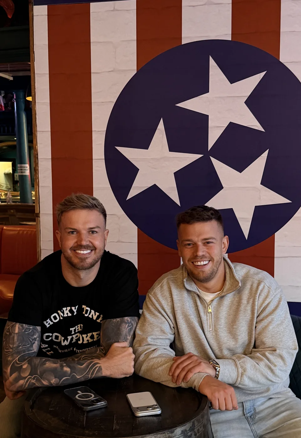 Ruby Rae's co-owners Dean Saunders, left, and Dan Cooke sitting at a barrel table in front of a Tennessee flag mural