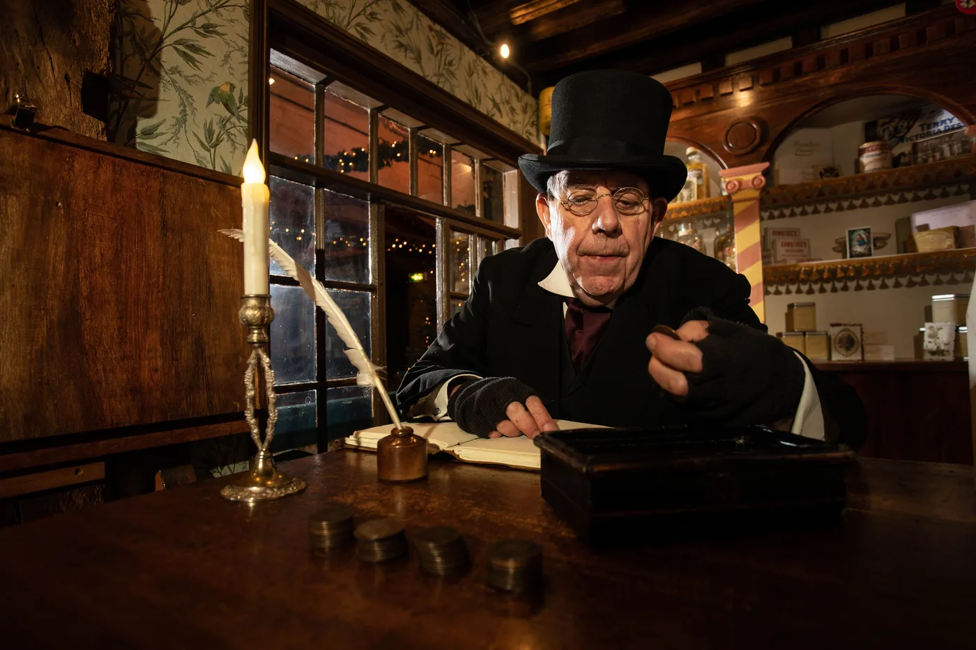 Scrooge in a top hat at a candlelit desk with stacks of coins