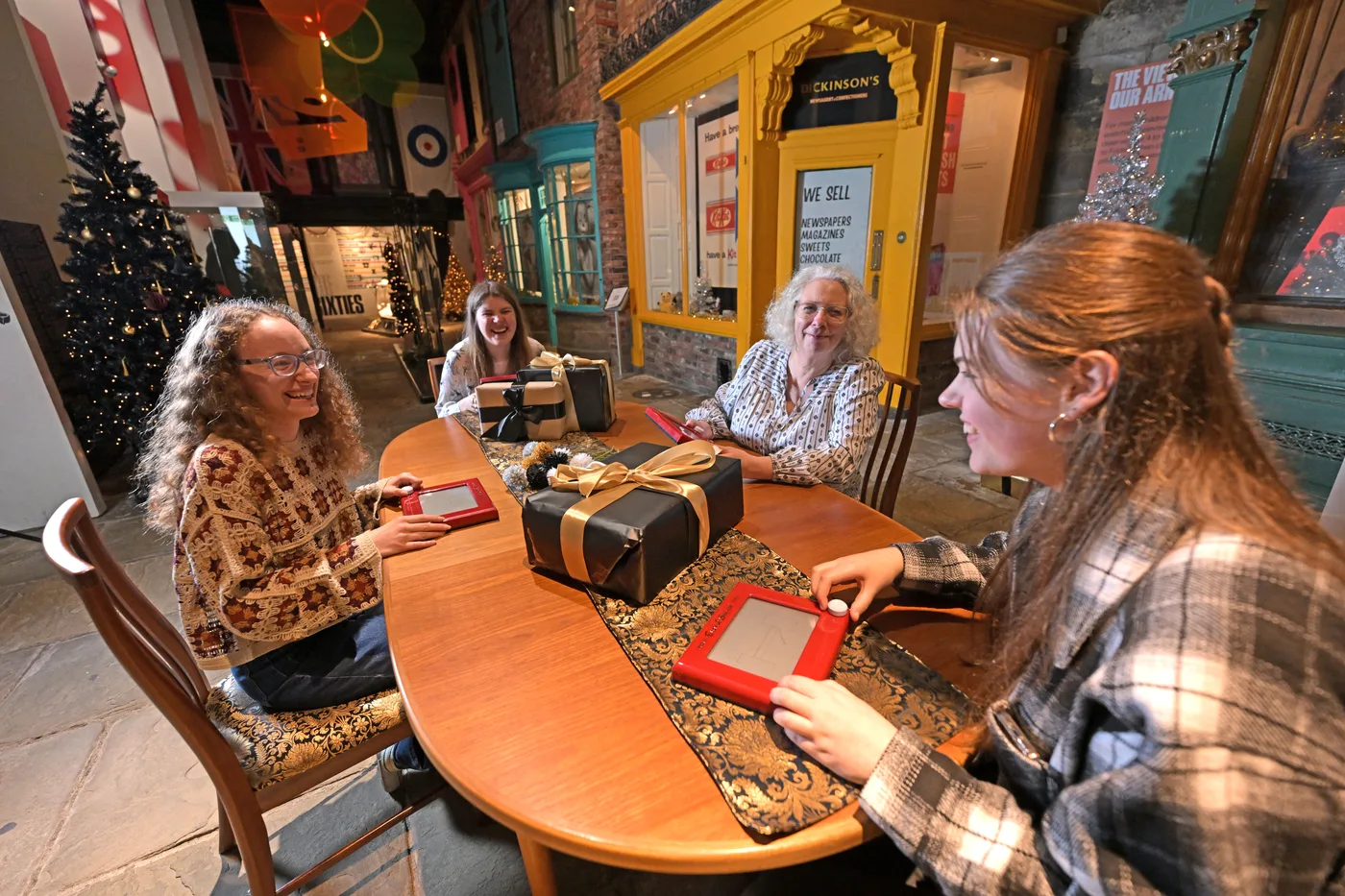 Four visitors around a table of wrapped presents on the Sixties street, with shop fronts behind