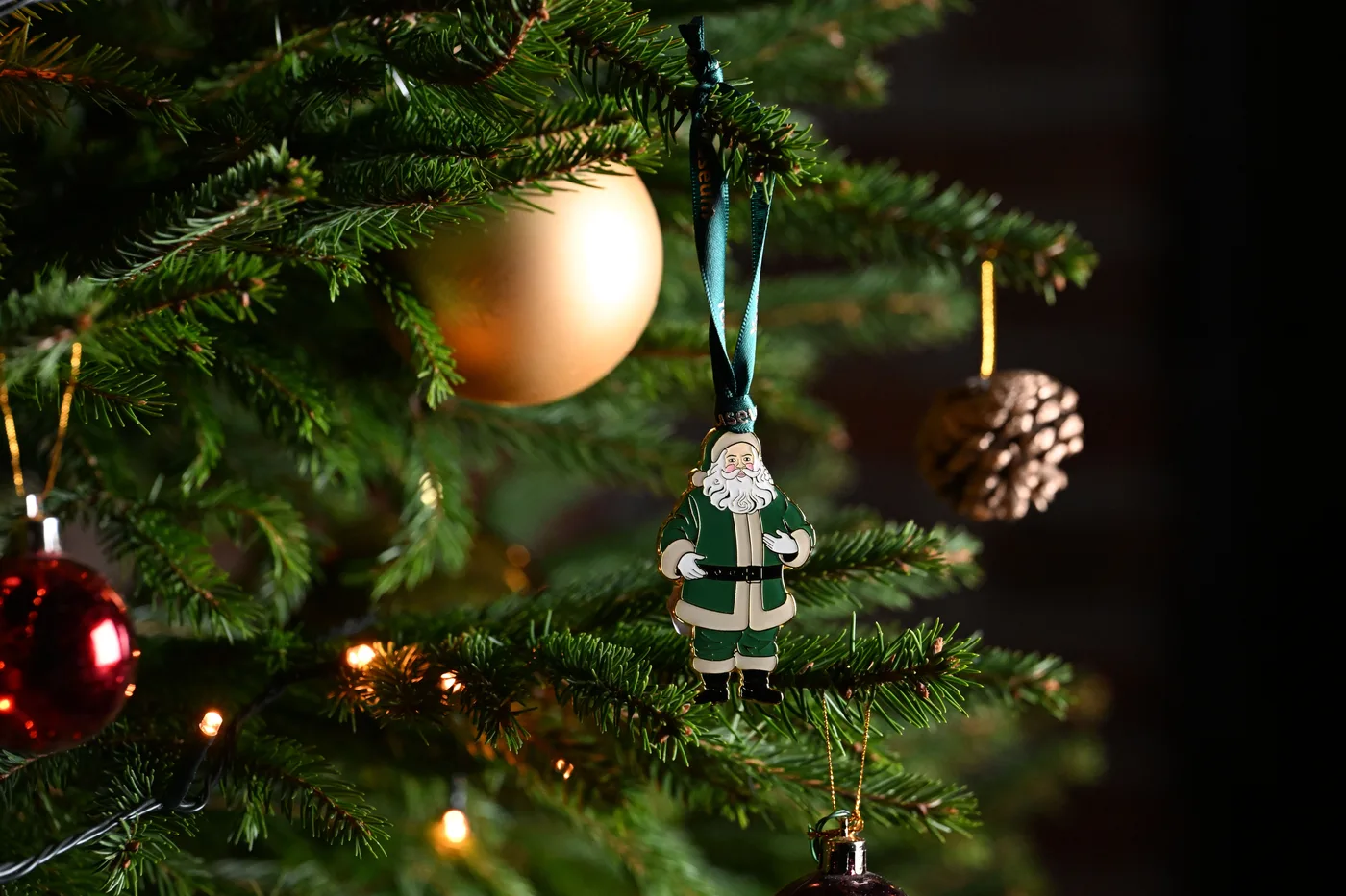 A green Father Christmas hanging decoration on a Christmas tree beside a gold bauble and a pine cone