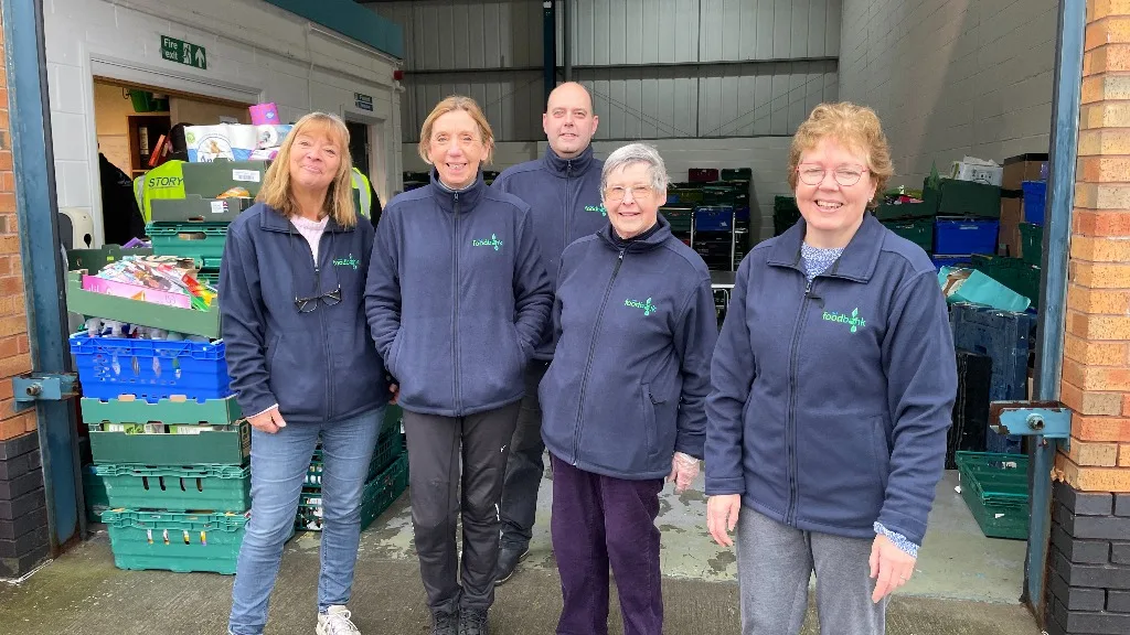 Five members of the York Foodbank team in navy fleeces standing among stacked crates of food