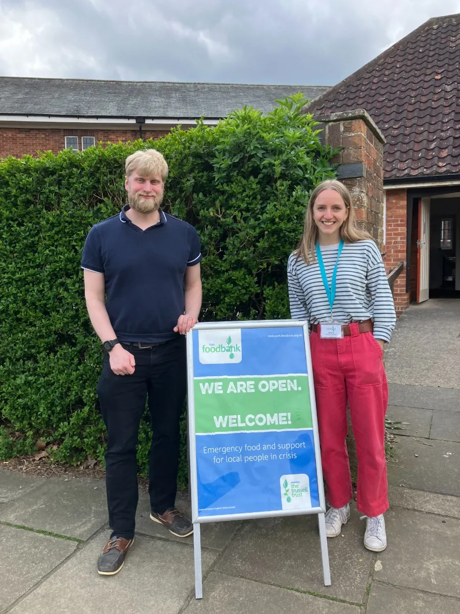 Two people standing outside a brick church hall beside an A-board sign reading We are open, welcome