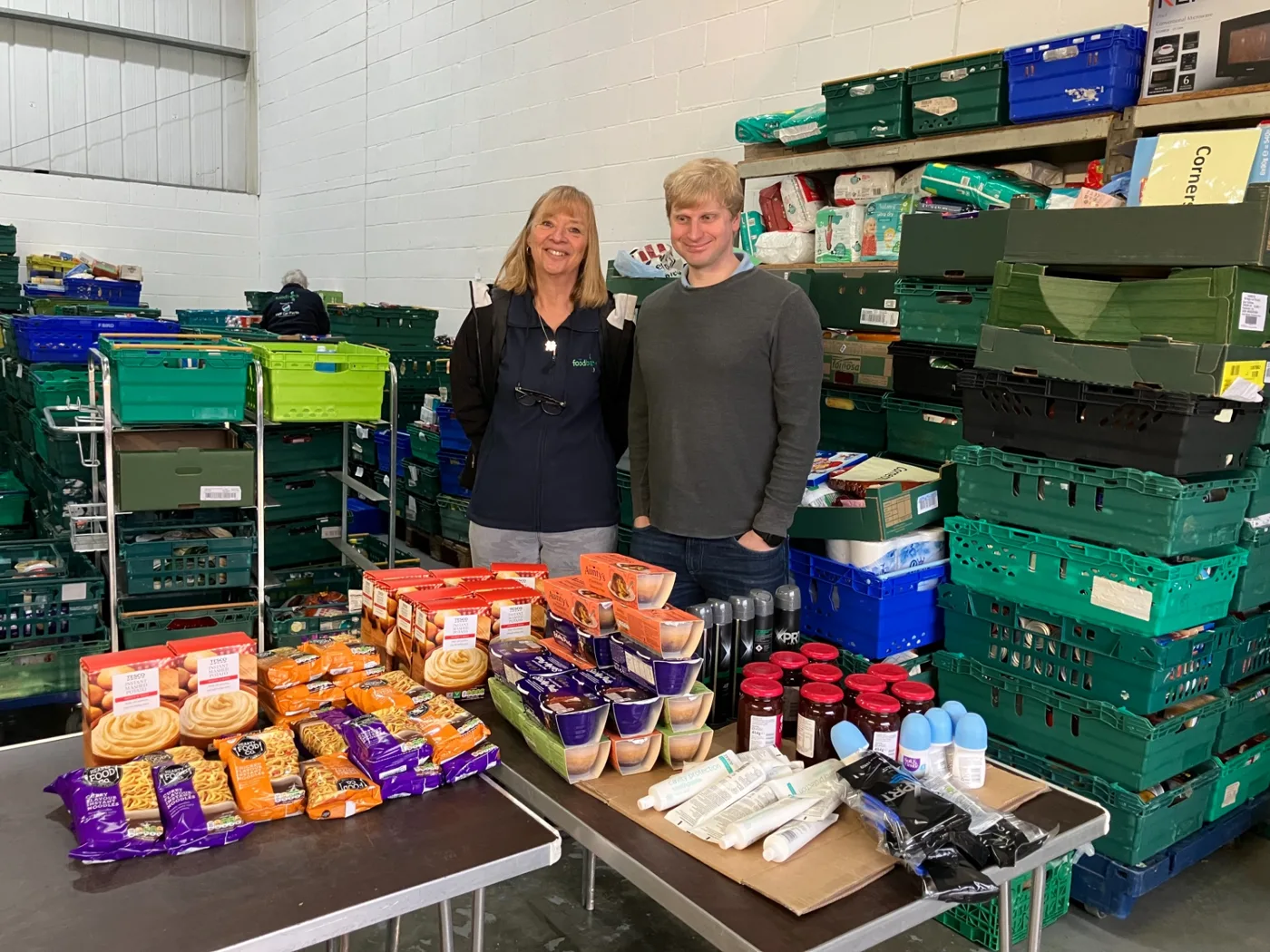 Two members of the York Foodbank team in a warehouse behind a table laid with donated food, crates stacked on shelves behind them