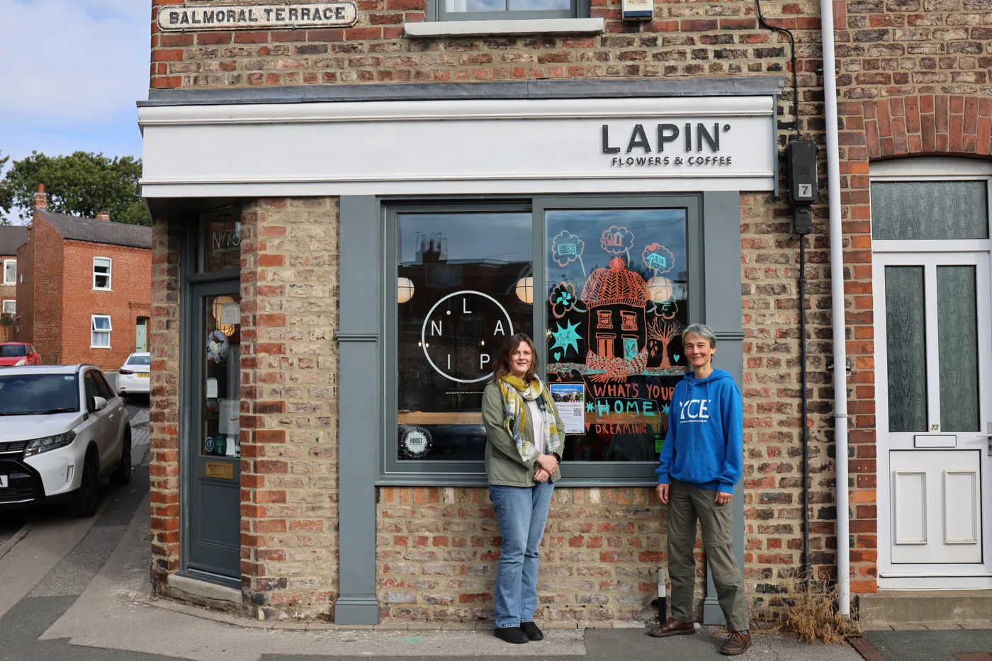 Two women standing outside Lapin Flowers and Coffee on Balmoral Terrace, York, beside a window painted with a house and the words What's your home dreaming