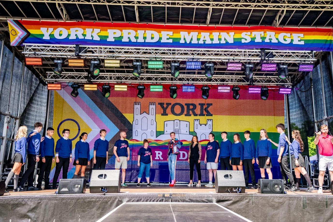 Young performers in school uniform and cast members in matching T-shirts lined up across the York Pride main stage, under a rainbow York Pride Main Stage banner