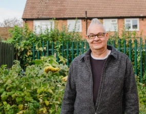Kevin, a smiling man with glasses and grey hair, standing in a community garden in front of a green fence and brick houses